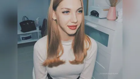 Smiling blonde woman in a white long-sleeve top sitting in a bright, modern minimalist room.