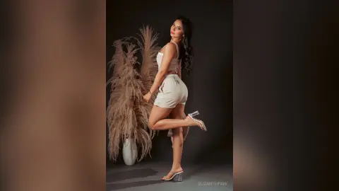 Elegant woman in white summer outfit posing with pampas grass in a moody studio setting.