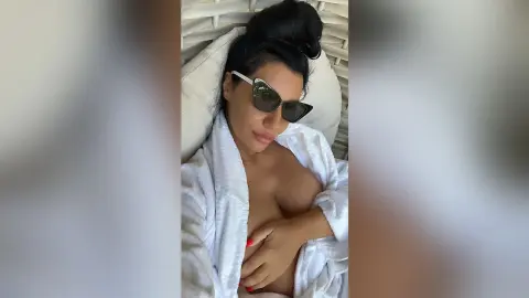 MissyJolie reclines against white pillows wearing a white robe and dark sunglasses, holding her hand against her bare breast.