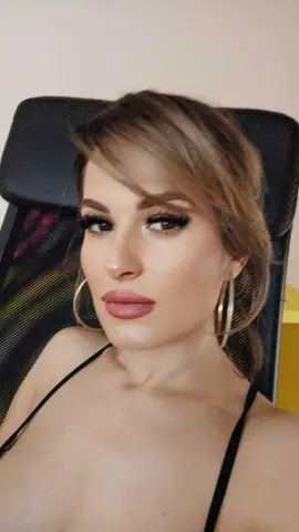 Glamorous woman with long lashes and gold hoop earrings wearing a black strap top in a cozy indoor setting.