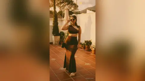 MiaBolson — Stylish woman in a black satin crop top and long skirt posing on a sunny outdoor terrace at sunset.