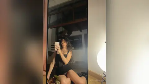 Young woman in a black mesh outfit taking a moody mirror selfie in a dimly lit modern room.