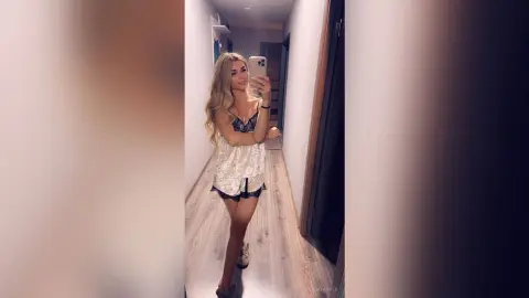 Blonde woman in a white lace nightgown taking a mirror selfie in a modern hallway.