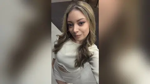 Smiling young woman in a white ribbed top posing for a selfie in a bright indoor setting.