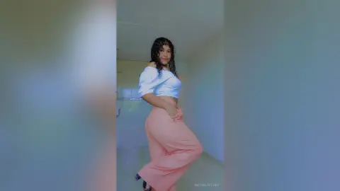 Young woman in a white off-the-shoulder top and peach trousers posing in a bright modern room.