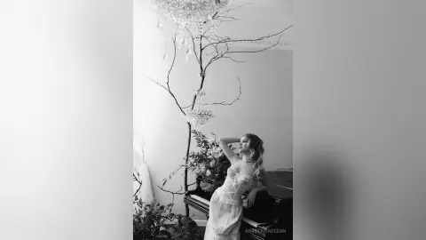 Elegant woman in a white lace gown posing gracefully under a decorative tree in a dreamy black and white setting.