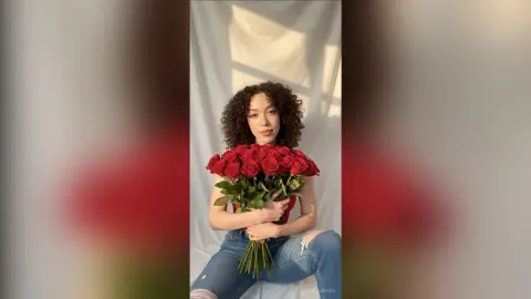 Young woman in a red top and denim jeans holding a large bouquet of red roses in a bright, minimalist setting.
