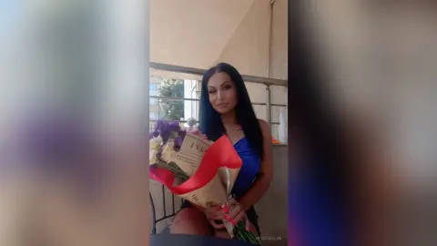 Beautiful woman in a blue top holding a large bouquet of flowers on a sunny balcony.