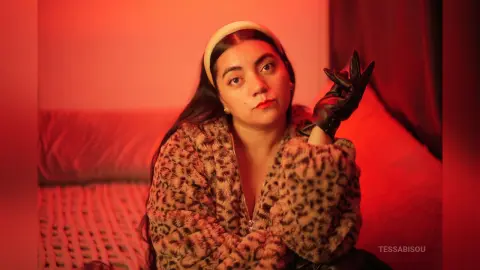 Young woman in leopard print coat and black gloves sitting on a sofa under warm red lighting.
