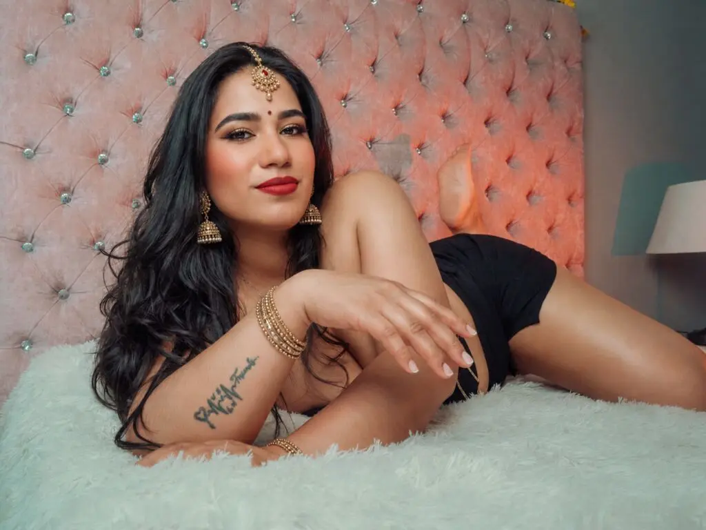Beautiful woman in black outfit and gold jewelry relaxing on a white bed against a pink tufted headboard.
