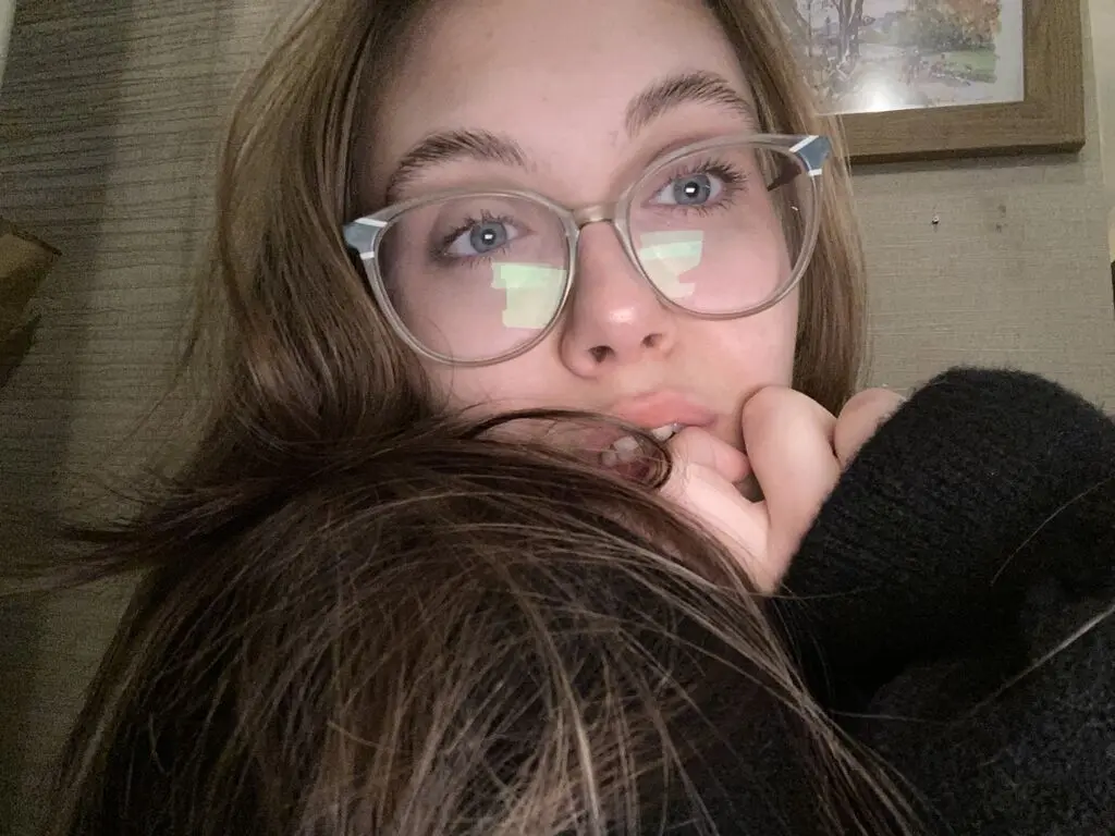 EmilyLoven wearing clear glasses and a black sweater, looking at the camera while biting her finger.