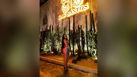 Stylish woman in a sleek dress posing at night in front of a glowing Dos Santos neon sign and tall cacti.