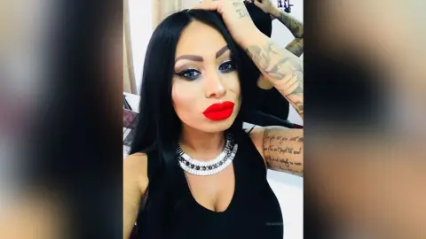 DeeaAnderson poses with her hand in her long black hair, wearing bright red lipstick and a black sleeveless top with a silver beaded necklace.