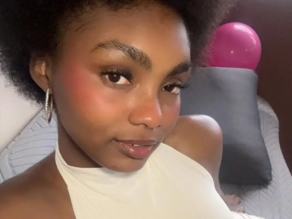 Close-up portrait of a smiling woman in a white halter top with natural makeup in a cozy indoor setting.