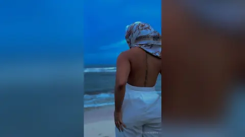 Woman in white linen outfit and headscarf facing a serene blue ocean at twilight.