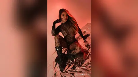 RosieThomas — RosieThomas poses in a sheer black patterned bodysuit and black gloves, sitting on a reflective silver foil background under red lighting.