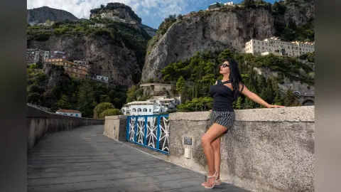 MissyJolie stands outdoors against a stone wall, wearing a black tank top and patterned shorts with her long dark hair down.