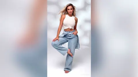 Blonde woman in a white tank top and light wash denim jeans posing against a bright white studio background.
