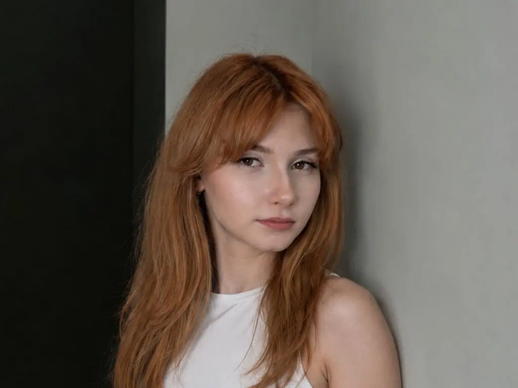 Portrait of a young woman with long red hair wearing a white top in a minimalist indoor setting.