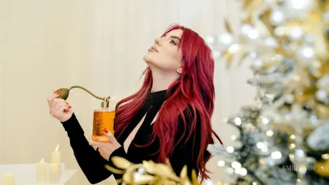 Woman with long red hair in a black dress holding a gold perfume bottle in a festive holiday setting.