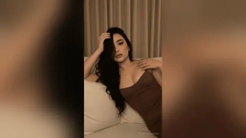 Elegant woman in a brown dress relaxing on a white bed in a cozy, warm-toned bedroom setting.
