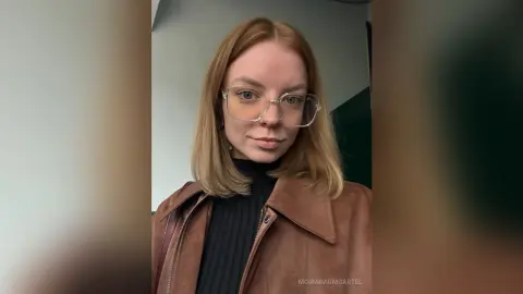 Redheaded woman in clear glasses and a brown suede jacket posing with a calm expression indoors.