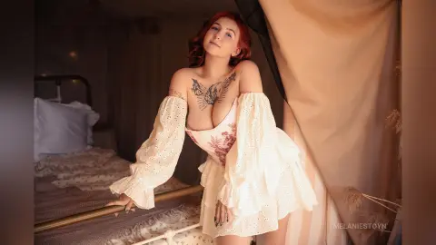 Redheaded woman in a white ruffled bohemian dress posing in a dreamy, sunlit bedroom setting.
