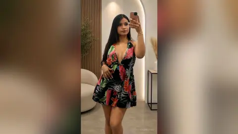 Young woman in a tropical floral print mini dress taking a mirror selfie in a modern minimalist room.