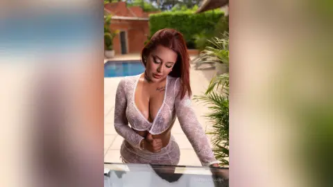 Elegant woman in a white lace long-sleeve dress posing outdoors by a swimming pool in soft sunlight.