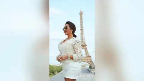 Dolce smiles in a white lace jacket and tight white skirt standing before the Eiffel Tower.