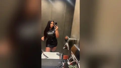 Young woman in a sleek black leather mini dress taking a mirror selfie in a modern bathroom.