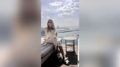 Stylish woman in a white crochet maxi dress sitting on a rooftop lounge overlooking a coastal city skyline.