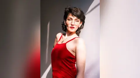IveteDurand wears a red tank top and glasses, posing with her dark hair styled up while looking toward the camera.