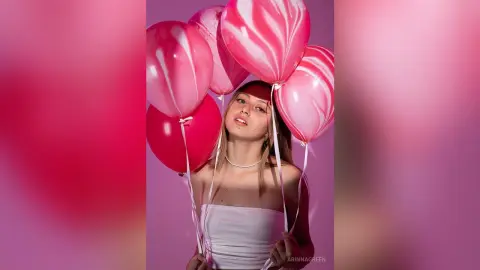 ArinnaGreen poses with pink and red balloons, wearing a white tube top against a pink background.