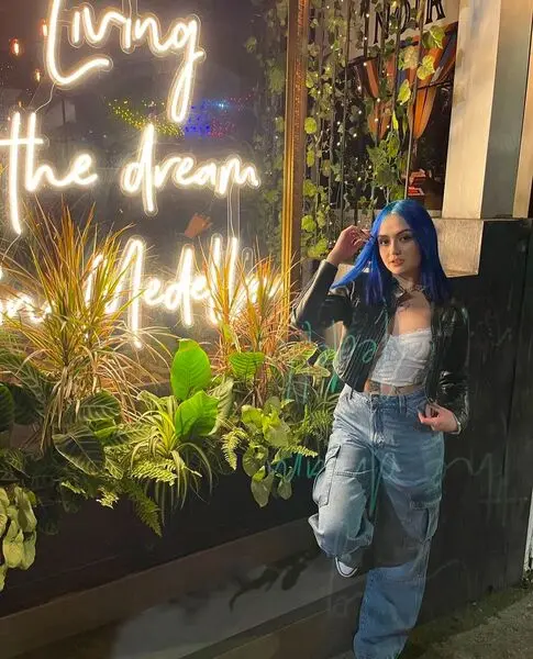 Andrea_Torner — Young woman with blue hair in a black leather jacket and denim posing by glowing neon signs at night.