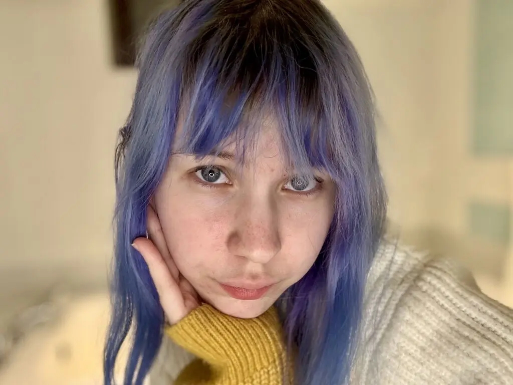 Young woman with blue hair and freckles wearing a cozy yellow sweater in a bright indoor setting.