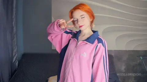 MozelleRozycki, a woman with bright orange hair, wearing a pink and navy blue track jacket while making a peace sign.