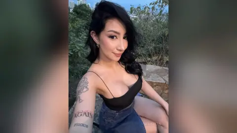 Smiling woman in a black bodysuit and denim shorts posing outdoors in a lush green garden setting.