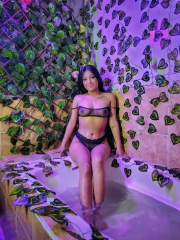 Young woman in black lace lingerie sitting in a bathtub surrounded by lush green ivy in moody purple lighting.