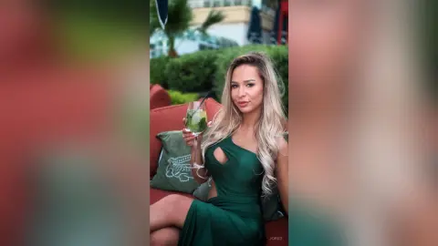 Smiling blonde woman in an elegant emerald green dress holding a cocktail at an outdoor lounge.