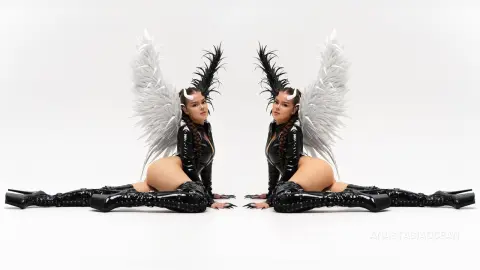 Woman in black latex bodysuit and feathered angel wings posing in a minimalist white studio setting.