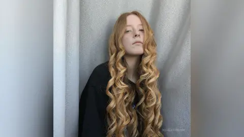 Redheaded woman with long wavy blonde hair wearing a black shirt in a minimalist grey studio setting.