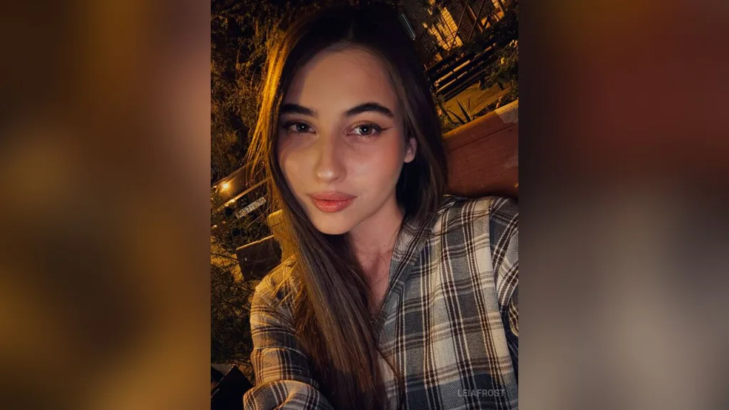 Young woman with winged eyeliner and plaid shirt smiling in warm nighttime outdoor lighting.