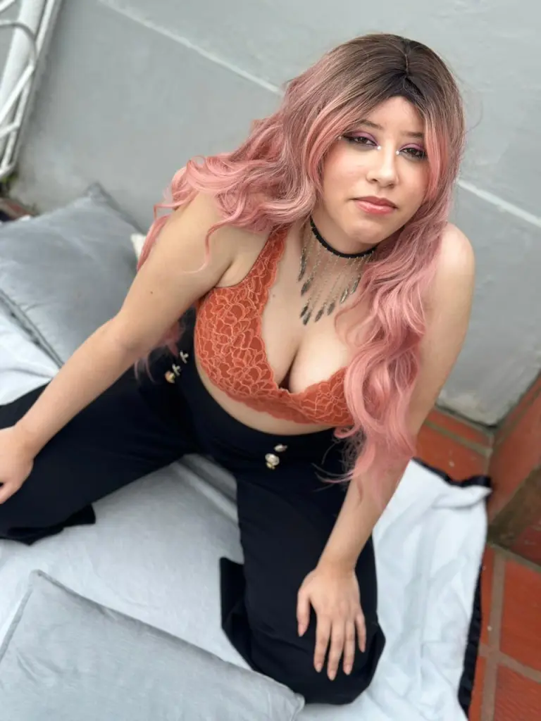 Izzumi smiles while leaning forward, wearing a pink wig and glasses, with her hands positioned near her chest.