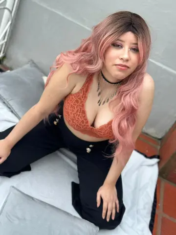Izzumi poses sitting on a light grey cushion wearing an orange lace bra and black trousers with her long pink hair falling over her shoulder.