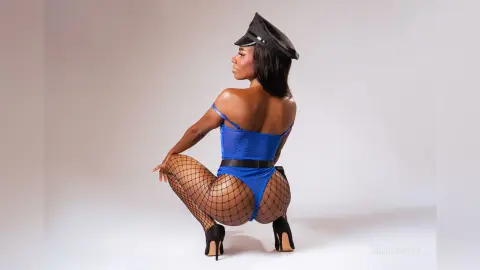 Black woman in blue bodysuit, fishnet stockings, and captain hat posing in a studio setting.