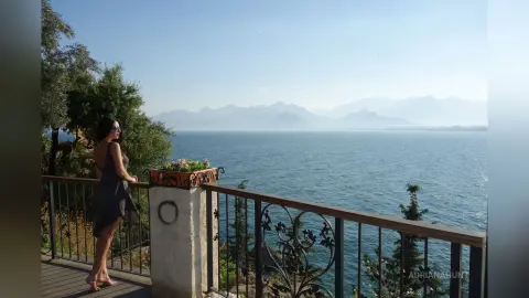 Woman in a black dress gazing at the scenic blue lake and mountains from a sunny balcony terrace.