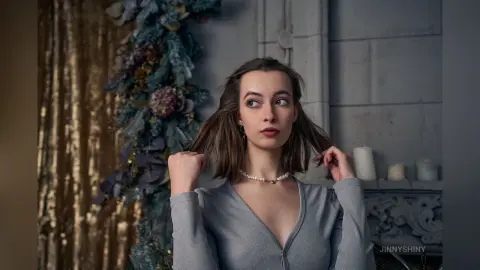 Young woman in a grey long-sleeve top posing thoughtfully in a moody, vintage-style room with floral decor.