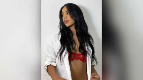 Young woman with long dark hair wearing a white button-down shirt and red lace bra in a bright indoor setting.