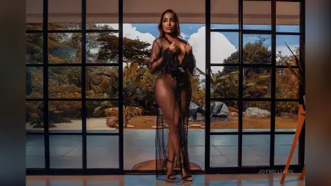 Elegant woman in a black sheer lace gown standing by large glass doors overlooking a lush tropical garden.